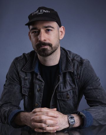 Rafael Fernández, a man with a short beard in a black cap and a dark denim jacket, hands folded, against a grey studio backdrop.